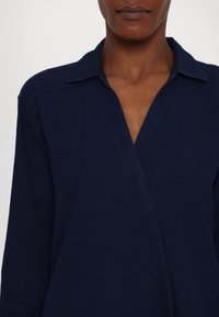 Femme portant une chemise bleu foncé à col avec un col en V profond et des manches longues sur un fond neutre.