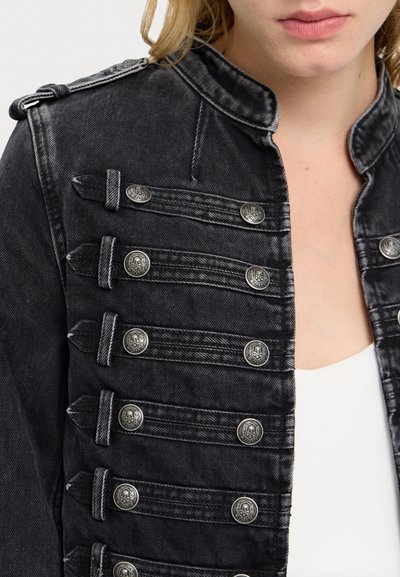 Nærbillede af en sort denim militærjakke med metalliske kranieknapper, båret over en hvid top.