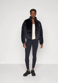 Sort jakke i imiteret pels med høj krave og lynlåslukning, med puffede sektioner. Båret over en beige top med sorte leggings og sneakers.