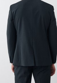 Chaqueta de traje a medida de color gris oscuro, con hombros estructurados, dos aberturas en la parte trasera y puños con botones, confeccionada en un tejido liso y texturizado.