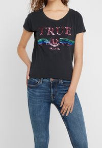 Woman wearing black t-shirt with pink sequined "TRUE" text and rainbow stripes, paired with blue jeans, posing with one hand in pocket.