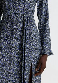 Blumenkleid mit schwarzem Hintergrund und blauen sowie weißen Blumen, langen Ärmeln mit Rüschensaum und einem seitlichen Schlitz. Weicher Stoff.