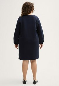 Dunkelblaues Kleid mit langen, puffigen Ärmeln und geradem Schnitt, ausgestattet mit einem runden Ausschnitt und glatter Textur. Kombiniert mit schwarzen Ballerinas.