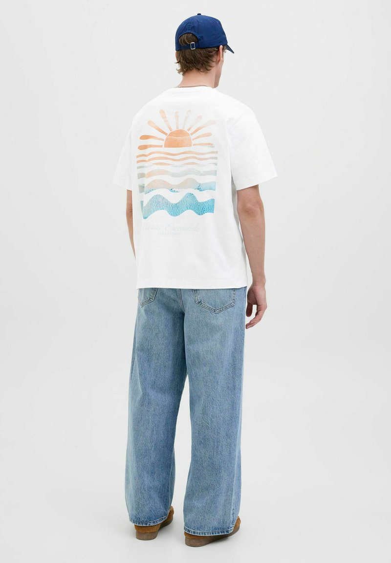 Witte katoenen t-shirt met een kleurrijke zonsondergang en golfafbeelding op de rug. Draagt het met lichtblauwe wijde jeans en een marineblauwe pet.