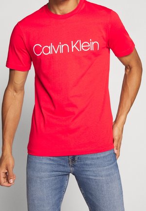 Muž v jasně červeném tričku Calvin Klein a modrých džínách stojí s jednou rukou v kapse na jednobarevném pozadí.