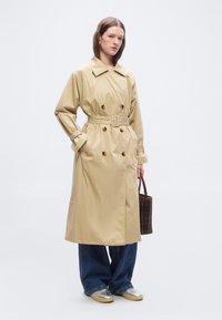 Femme portant un trench-coat beige ceinturé, un jean bleu à jambes larges, des baskets argentées et tenant un sac à main à carreaux foncés.