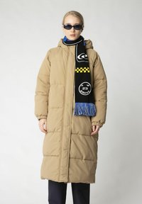 Beige puffer coat with a hood and elastic cuffs, featuring a zip closure. The model wears a black and yellow checkered scarf with blue fringes.
