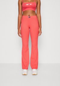 Kobieta nosząca koralowe, prążkowane spodnie dzwony i dopasowany krótki top z logo "Tommy Jeans", w połączeniu z białymi sneakersami.