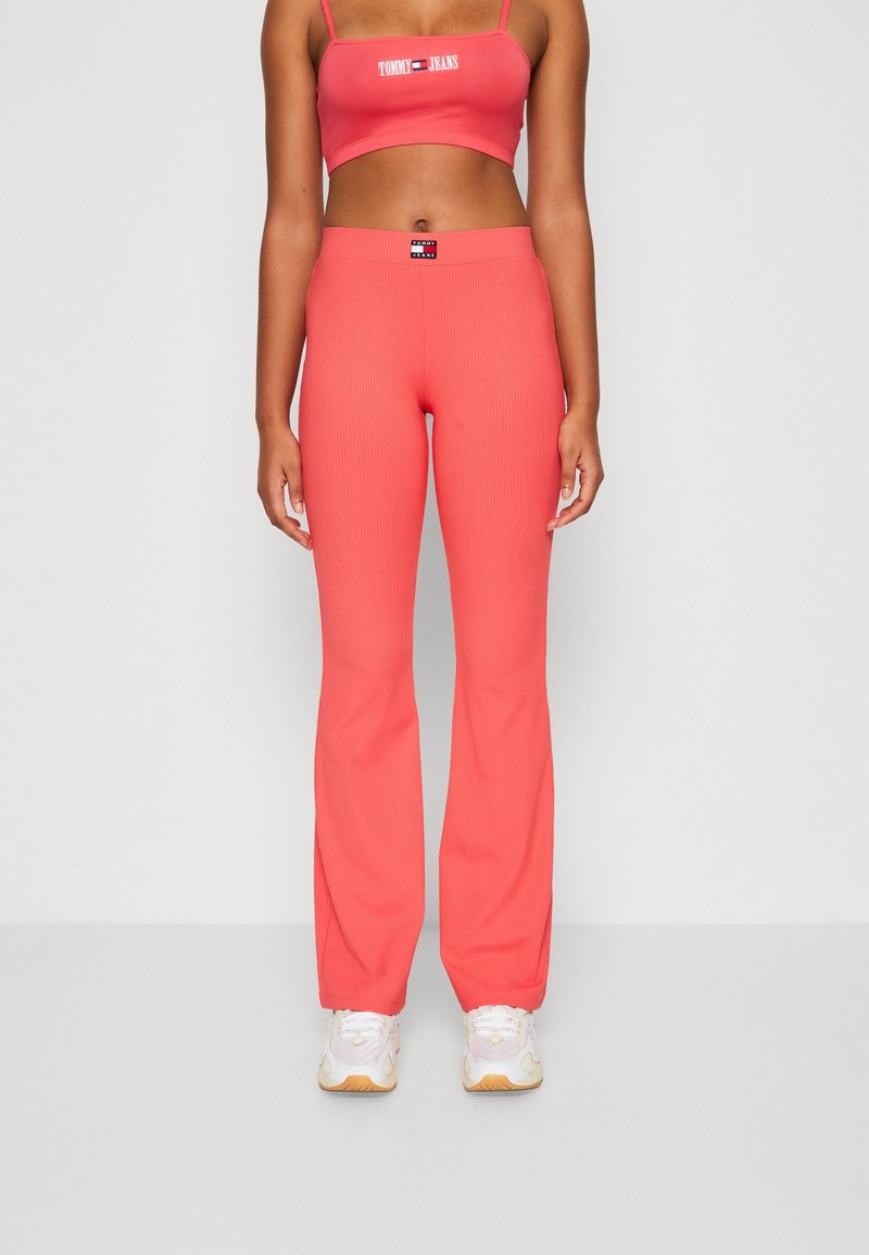 Kobieta nosząca koralowe, prążkowane spodnie dzwony i dopasowany krótki top z logo "Tommy Jeans", w połączeniu z białymi sneakersami.