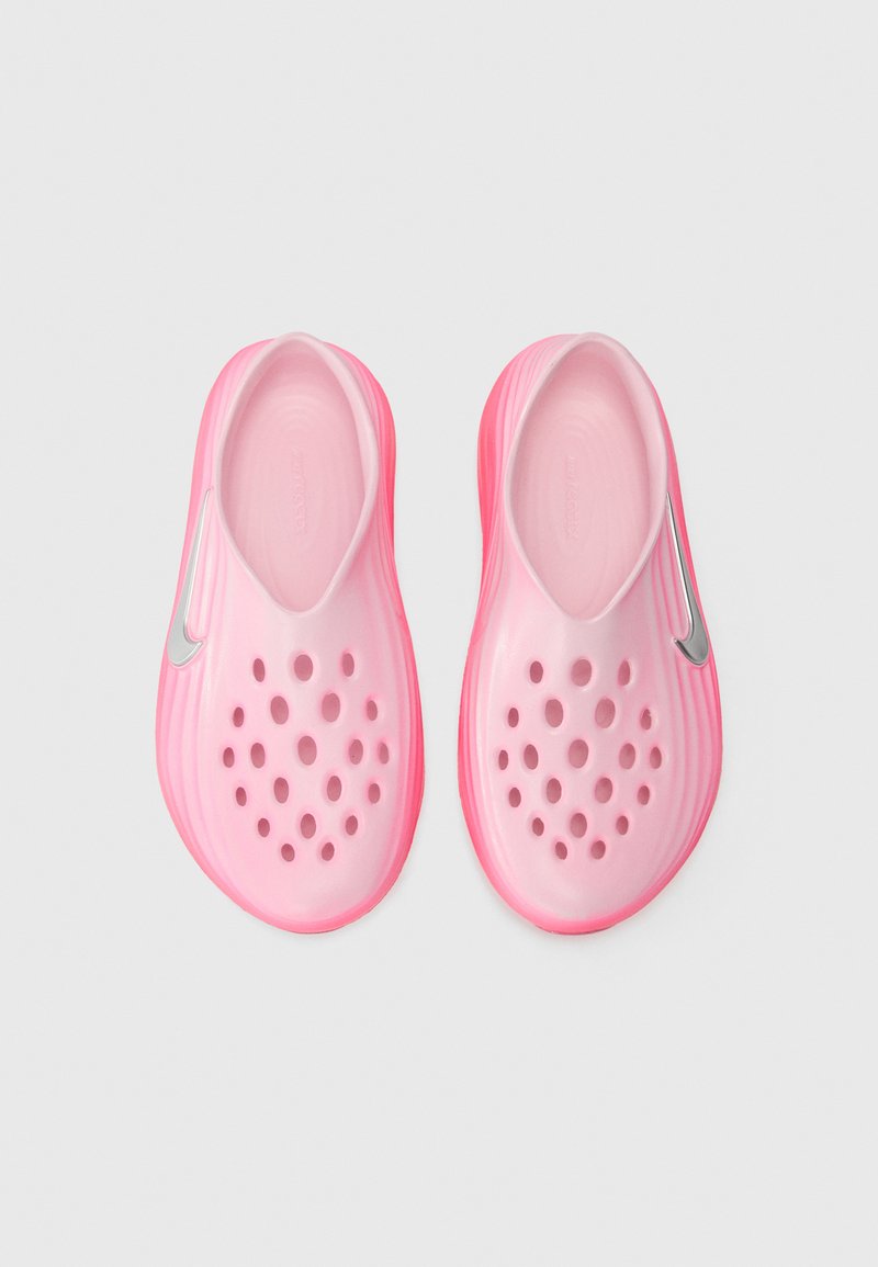 Zapatillas de color rosa sin cordones con una textura suave, que cuentan con múltiples agujeros para ventilación y un acento plateado en el lateral. Forma redondeada.