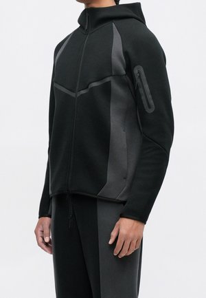 Personne portant une veste zippée noire et gris foncé avec capuche et pantalon assorti, debout devant un fond uni clair.