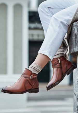 Legs in white pants wearing beige socks and brown leather ankle boots with buckle and zipper detail, sitting on stone ledge outdoors.