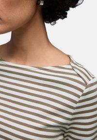 Frau trägt ein olivgrün-weiß gestreiftes Bootskragenhemd, zeigt Hals, Schulter und dunkles lockiges Haar mit silbernem Ohrring.