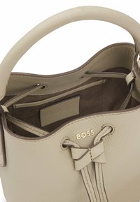 Beige leren bucketbag met een gestructureerde afwerking, trekkoordsluiting en een kleine strik als accent. Heeft een goudkleurig logo aan de binnenkant.