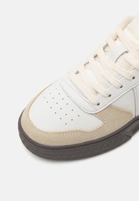 White leather sneaker with beige suede accents, round toe, perforated detailing, and cream laces. Textured dark rubber outsole.