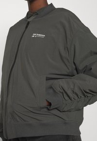 Giacca bomber grigia con zip frontale, polsini e colletto a costine, e un piccolo logo bianco "New Balance" sul petto.