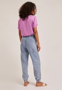 Žena s kudrnatými vlasy, oblečená v růžové blůze s krátkým rukávem, světle modrých džínách s elastickým pasem a béžových sandálech, stojící na jednobarevném pozadí.