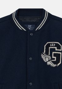 Marineblaue Collegejacke mit gestreiftem Kragen, Druckknöpfen und einem Aufnäher mit dem Buchstaben G, Flügeln und einem Laufschuh auf der Brust.