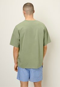 Olivza zaļa t-krekls ar īsām piedurknēm un brīvu piegriezumu, kopā ar ziliem un baltiem svītrainiem peldkostīmiem, redzams no aizmugures.