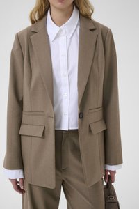 Brun skræddersyet blazer med to forlommer, hakkede revers og enkelt knap. Underneden en hvid skjorte med krave og synlige knapper.