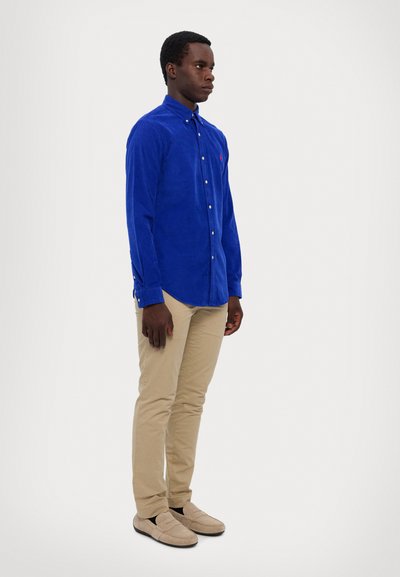 Cămașă din catifea cu un design cu nasturi, cu mâneci lungi și un buzunar pe piept, asortată cu pantaloni slim-fit bej și încălțăminte de culoare deschisă.