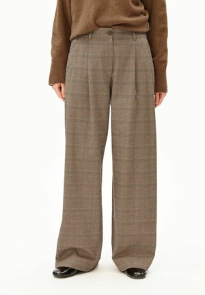 Personne portant un pantalon large taille haute marron à carreaux avec des plis devant, un pull marron et des chaussures noires, debout sur un fond blanc.