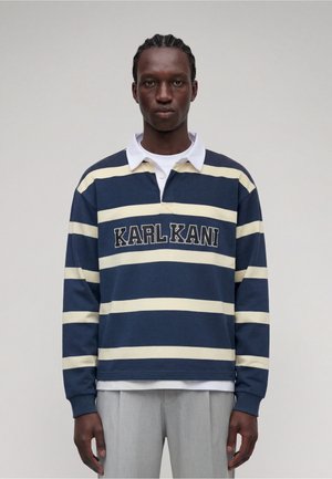 Homme aux cheveux tressés portant un maillot de rugby rayé bleu marine et crème avec le texte "KARLKANI", debout devant un fond clair uni.