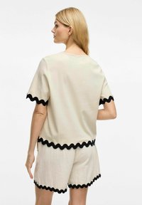 Crème-kleurig top met korte mouwen en zwarte golvende rand; gecombineerd met bijpassende crème shorts. Gemakkelijke stof, relaxte pasvorm en minimalistisch ontwerp.