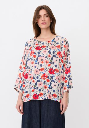 Femme portant un chemisier ample à fleurs rouges, roses et bleues et un pantalon large foncé, debout devant un fond clair uni.