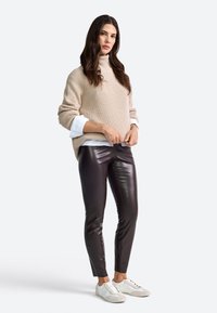Maglione a collo alto a coste beige indossato sopra una camicia bianca, abbinato a pantaloni aderenti in eco-pelle marroni e sneakers bianche.