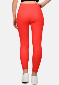 Rode legging met een hoge tailleband, gemaakt van rekbaar materiaal. Soepele textuur zonder patronen. G gedragen met witte sportschoenen.