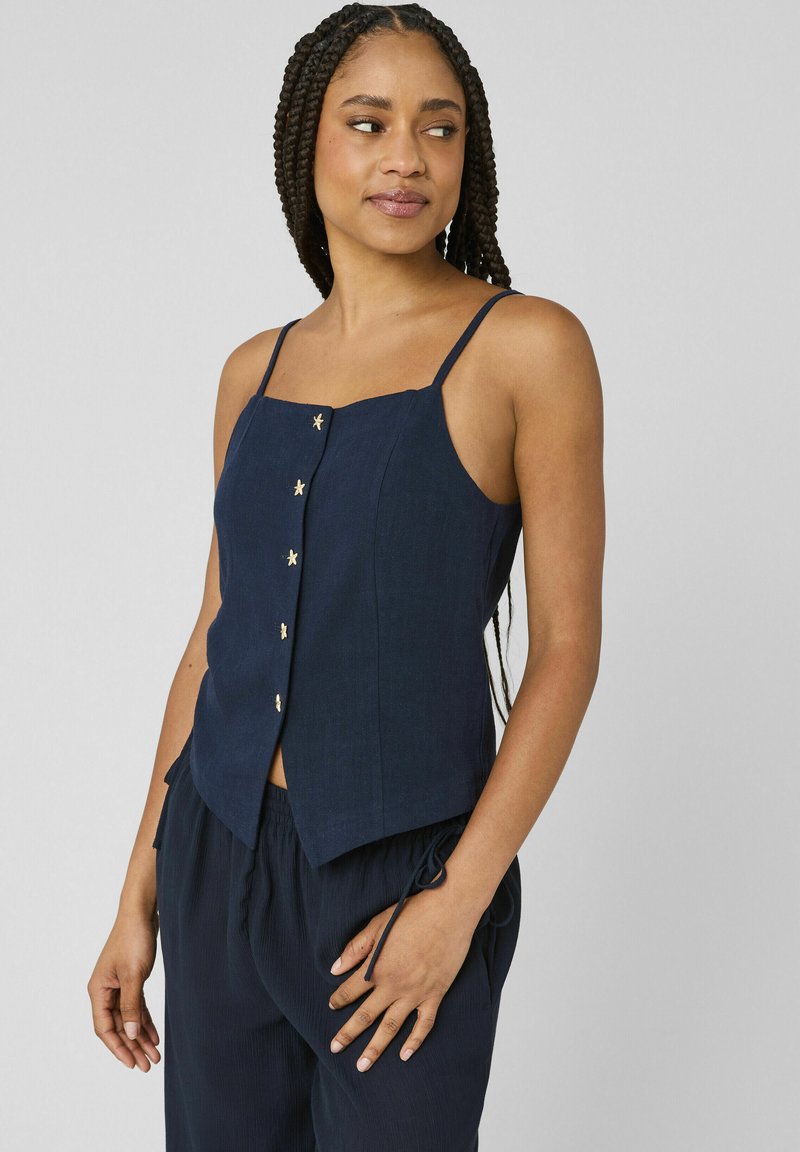Femme aux cheveux tressés portant un haut sans manches bleu marine avec des boutons en forme d'étoile et un pantalon ample assorti, debout devant un fond uni.