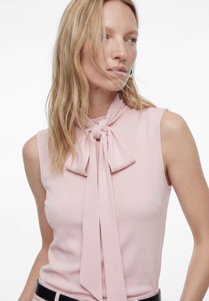 Femme aux cheveux blonds portant un haut rose clair sans manches avec un nœud noué au niveau du cou, regardant sur le côté sur fond blanc.