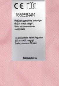 Etikett på en rosa tygbakgrund. Vit tagg med säkerhetsinformation, CE-märke och text som anger överensstämmelse med PPE-regler.