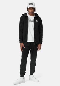 Sort hoodie og joggingsbukser med hvidt logo, parret med en hvid t-shirt og kasket. Hvide sneakers fuldender outfittet. Casual atletisk stil.