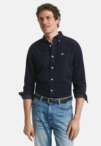 Marinblå kordskjorta med knapp-ner krage, vita knappar, broderad logotyp på bröstet; kombinerad med ljusblå denimjeans och svart vävd bälte.