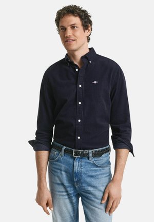 Marineblaues Cordhemd mit Button-Down-Kragen, weißen Knöpfen, gesticktem Logo auf der Brust; kombiniert mit hellblauen Denim-Jeans und einem schwarzen gewebten Gürtel.
