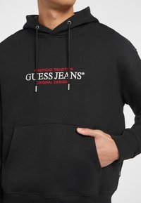 Svart hettegenser med snorer, frontlomme og brodert "GUESS JEANS"-logo i rødt og hvitt. Mykt materiale, klassisk passform.