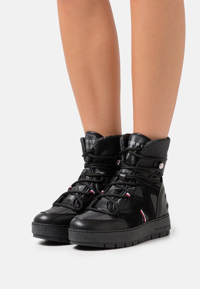 Baskets montantes noires brillantes avec semelles épaisses, col rembourré, lacets en corde, et accents du logo Tommy Hilfiger portées sur des jambes nues.