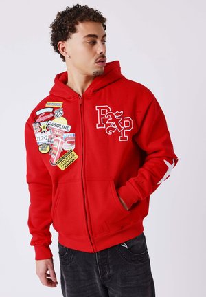 Homme portant un sweat à capuche rouge zippé avec des patchs colorés sur le thème de l'essence et un motif d'étoile blanche sur la manche, regardant vers sa gauche.