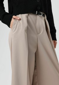 Beige wijde broek met plooien aan de voorkant, gemaakt van een gladde stof. Zwarte geribbelde cardigan met grote knopen en een zilveren gespdetail.