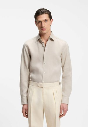 Mann mit zurückgekämmt Haar, der ein beiges, langärmliges Hemd mit Knöpfen trägt, das in hoch taillierte, cremefarbene, plissierte Hose gesteckt ist, vor einem schlichten Hintergrund.
