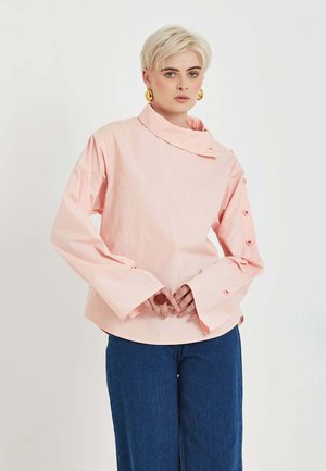 Osoba s krátkymi blond vlasmi, ktorá má na sebe nadrozmerný svetloružový dlhý rukáv s detailmi na gombíky na rukávoch a modré nohavice, stojaca na jednoliatom pozadí.