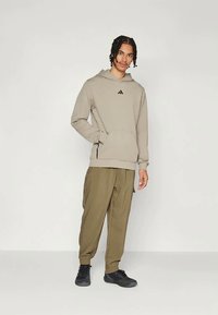 Bežs kapuce krekls ar priekšējo kabatu un melnu logo, apvienots ar oliņzaļa brīva piegriezuma biksēm un melnām kurpēm, uz neitrāla fona.