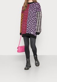 Bunter, übergroßer Strickpullover im Schachbrettmuster, mit lila, pinken, orangen und schwarzen Abschnitten; kombiniert mit schwarzer Jeans und Kampfstiefeln.