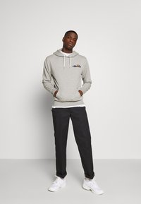 Sudadera gris con bolsillo frontal, cordones blancos y logotipo bordado. Combinada con pantalones negros y zapatillas blancas. Textura de tela suave.