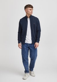 Granatowa kurtka zapinana na zamek z prążkowaną fakturą, wysokim kołnierzem oraz naszywką z logo. Noszona na białej koszulce, w połączeniu z niebieskimi dżinsami i jasnymi trampkami.