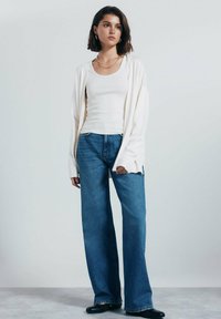 Bílý dlouhý svetr s dlouhým rukávem přes bílou tílko, spárovaný s volnými modrými džínami a černými nazouvacími botami. Jednoduchý design, bez výrazných vzorů.