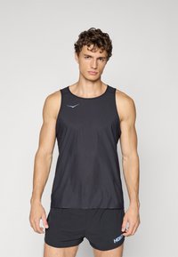 Mann in schwarzem ärmellosem Sportshirt und schwarzen Shorts, steht vor einem hellgrauen Hintergrund, schaut nach vorne mit neutralem Ausdruck.
