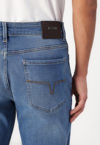 Jeans din denim albastru, cu un buzunar posterior cu un model de cusătură distinctiv și o etichetă din piele neagră la brâu cu inscripția "JOOP! JEANS" embosată.